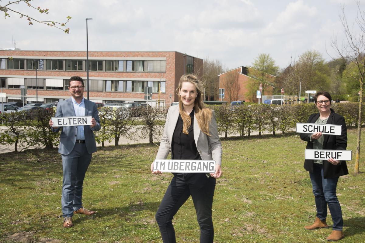Projekt "Mein Kind - im Übergang Schule und Beruf" Drei Personen stehen auf einer Wiese vor einem Bürogebäude und haben Schilder in den Händen.