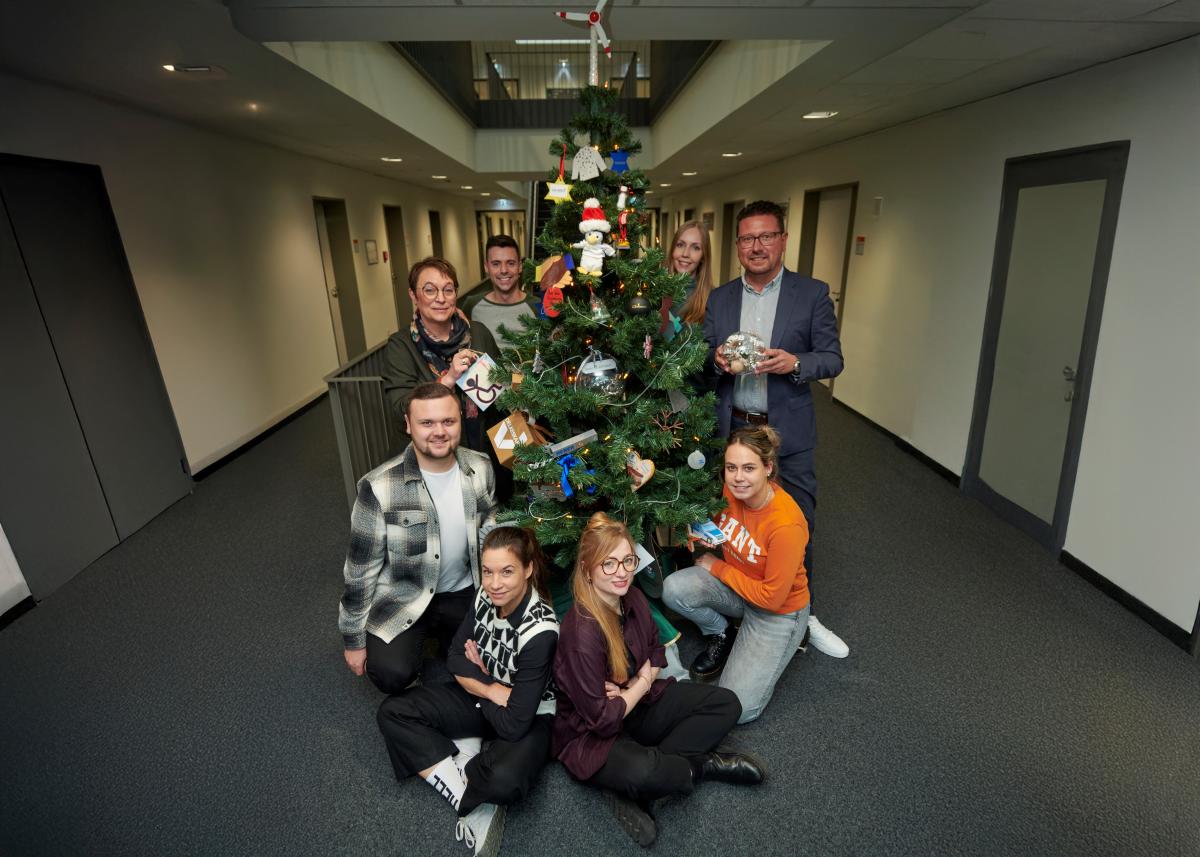 Eine Personengruppe steht um einen Weihnachtsbaum in einem Flur herum.