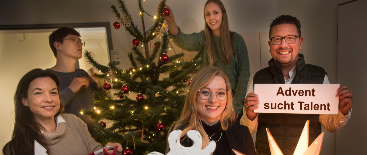 Eine Personengruppe steht um einen Weihnachtsbaum herum und hält Weihnachtsdekoration in den Händen.