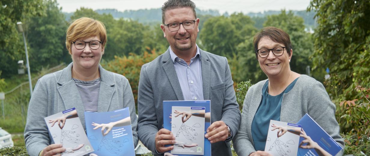 Drei Personen mit Broschüren in den Händen stehen auf einer Dachterrasse.