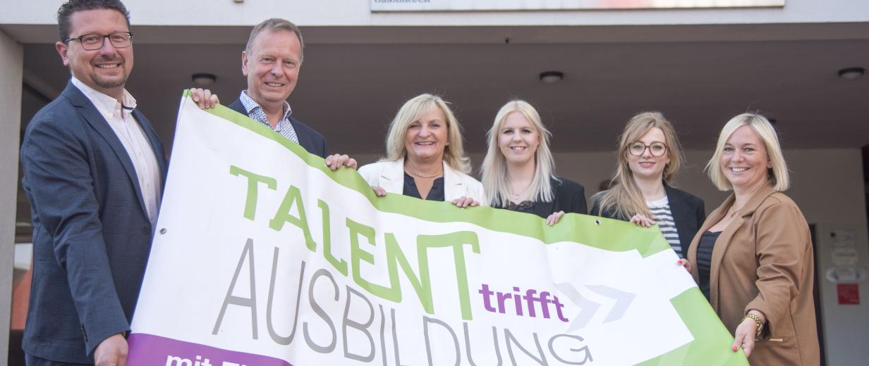 Sechs Personen mit einem Werbebanner in den Händen stehen vor einem Schulgebäude.