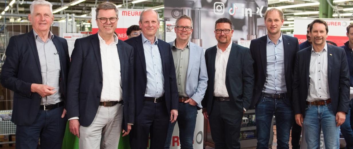 Sieben Personen stehen in einer Werkhalle.