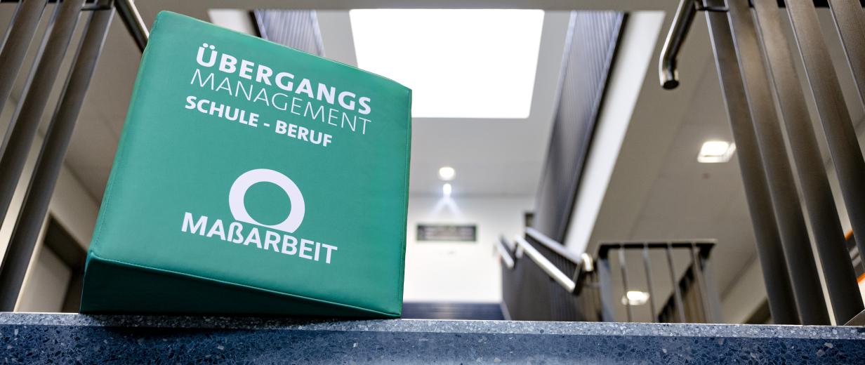 Ein großer Würfel mit MaßArbeit Logo liegt auf einer Treppe.