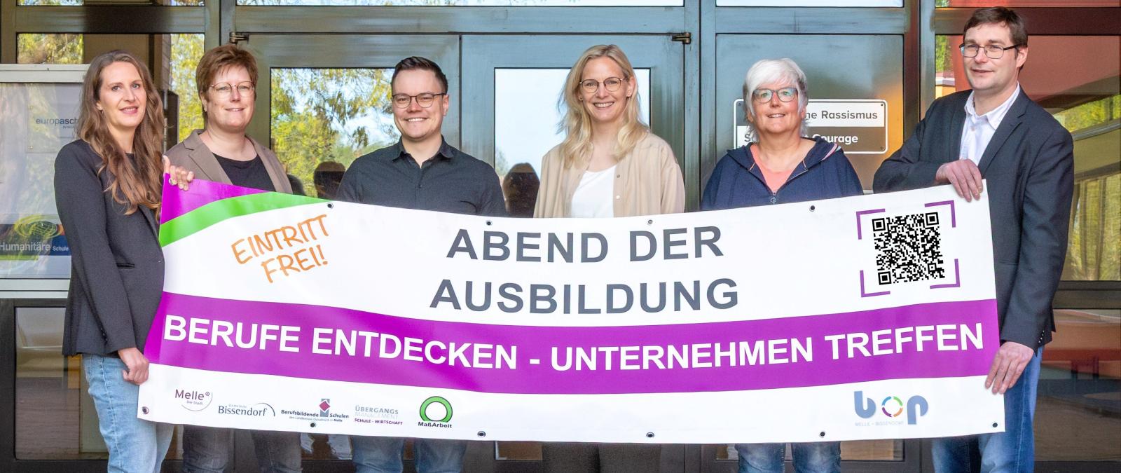 Sechs Personen mit einem Werbebanner in den Händen stehen vor einem Gebäudeeingang.