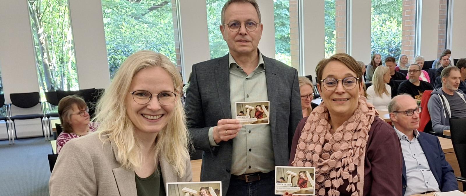 Drei Personen in einem Versammlungssaal halten Flyer in die Kamera.
