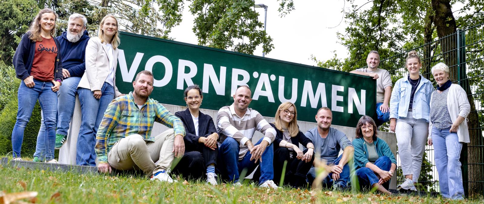 Eine Personengruppe steht, hockt und sitzt vor einem Zaun.