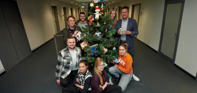 Eine Personengruppe steht um einen Weihnachtsbaum in einem Flur herum.