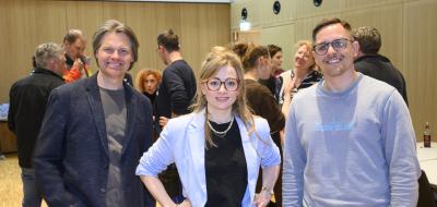 Drei Personen stehen mit anderen in einem Raum und schauen in die Kamera.