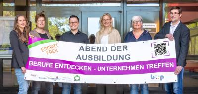 Sechs Personen mit einem Werbebanner in den Händen stehen vor einem Gebäudeeingang.