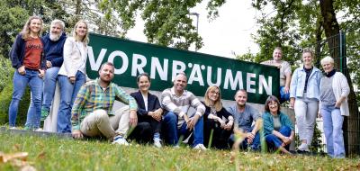 Eine Personengruppe steht, hockt und sitzt vor einem Zaun.