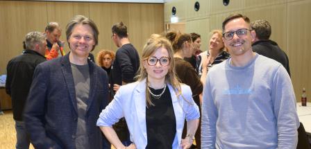 Drei Personen stehen mit anderen in einem Raum und schauen in die Kamera.