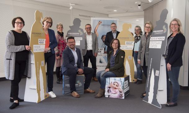 Eine Personengruppe steht in einem Raum zwischen Aufstellern, die menschliche Silhouetten darstellen.