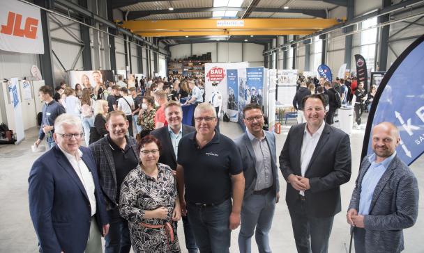 Eine Personengruppe steht in einer Werkhalle, im Hintergrund sind Messestände und Besucher zu sehen.