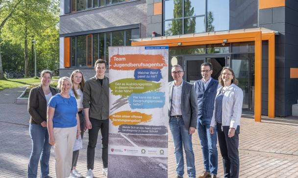 Sieben Personen stehen vor einem Schulgebäude in ihrer Mitte ein Rollup-Poster.