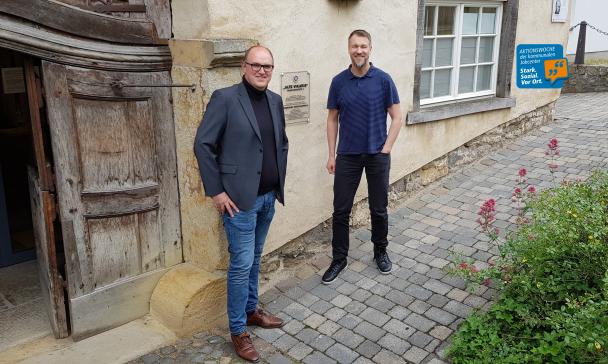 Zwei Personen stehen vor einem historischen Gebäude auf einem gepflasterten Weg.