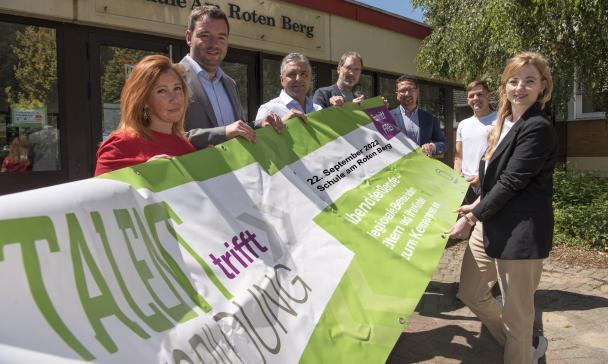 Sieben Personen stehen vor einem Schulgebäude und halten ein Werbebanner in ihren Händen.