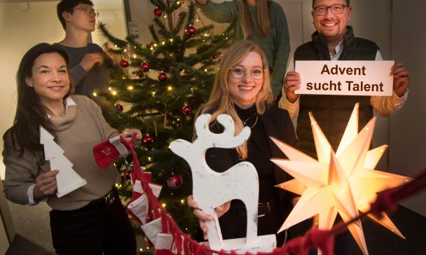 Eine Personengruppe steht um einen Weihnachtsbaum herum und hält Weihnachtsdekoration in den Händen.
