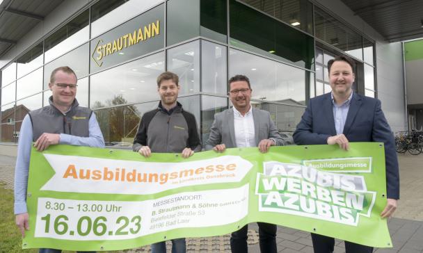 Vier Personen stehen vor einem Firmengebäude mit Glasfassade und halten ein Banner in den Händen.