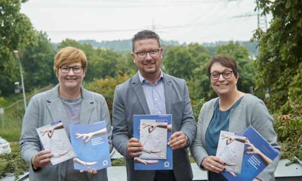 Drei Personen mit Broschüren in den Händen stehen auf einer Dachterrasse.