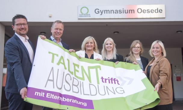 Sechs Personen mit einem Werbebanner in den Händen stehen vor einem Schulgebäude.