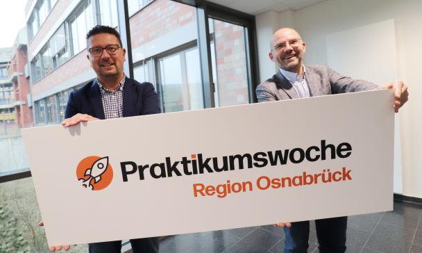 Zwei Personen mit einem Schild in den Händen stehen in einem Gebäudeflur.