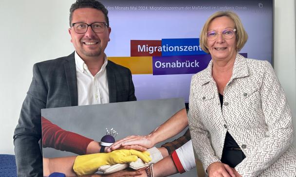 Zwei Personen mit einem großen Foto in den Händen stehen vor einem Display.