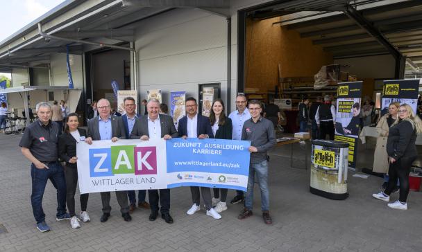 Eine Personengruppe mit einem Banner in den Händen steht vor einer Lagerhalle.