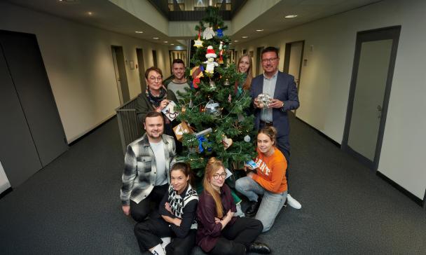 Eine Personengruppe steht um einen Weihnachtsbaum in einem Flur herum.