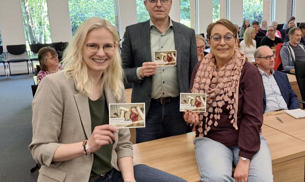 Drei Personen in einem Versammlungssaal halten Flyer in die Kamera.