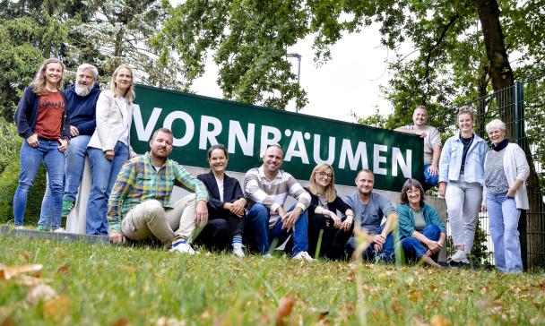 Eine Personengruppe steht, hockt und sitzt vor einem Zaun.